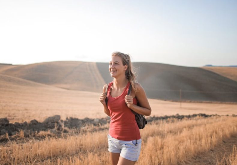Smiling woman in casual attire enjoying a summer hike through golden fields at sunset.